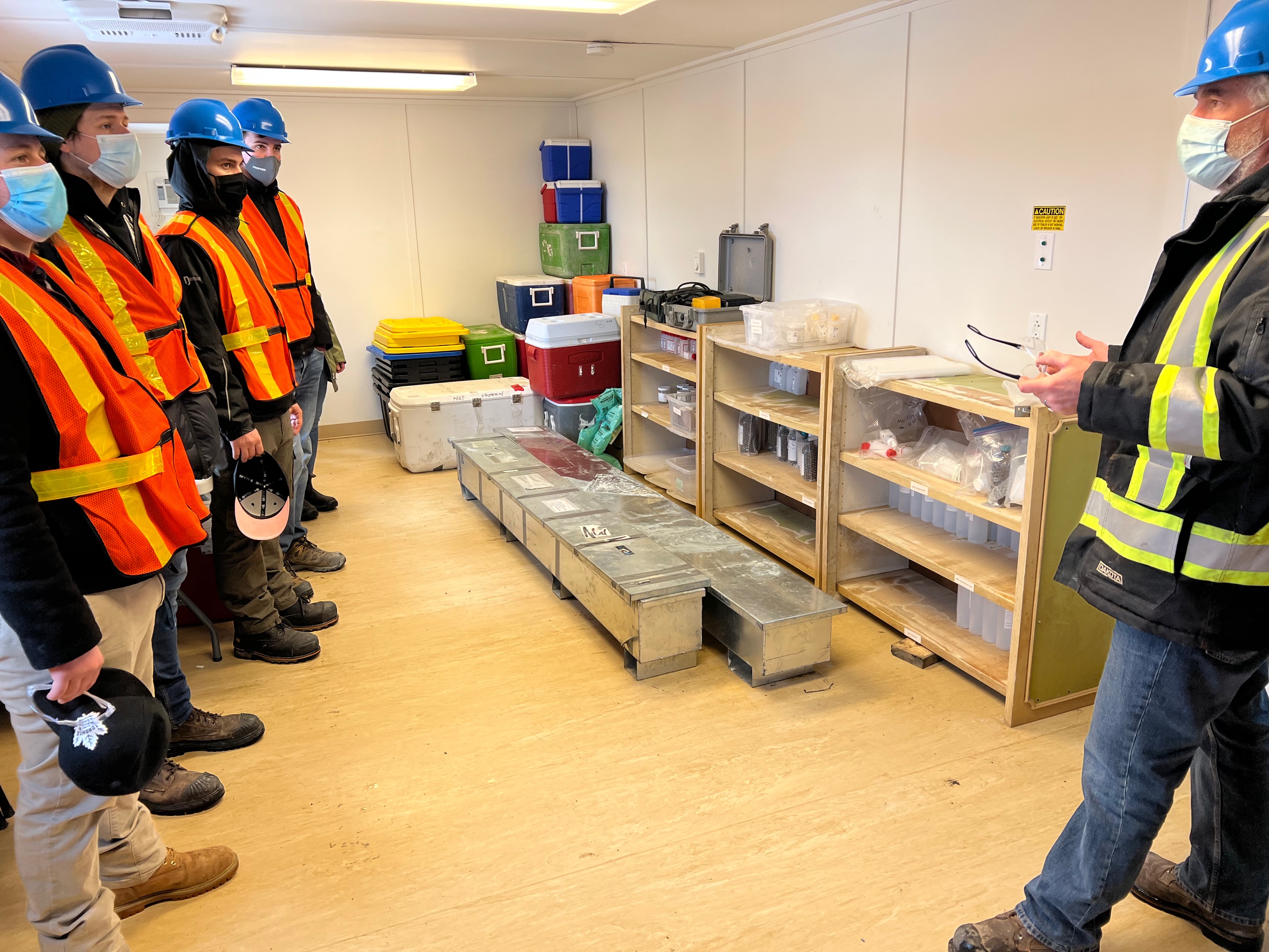 three people in safety vests and hard hats look at another person explaining what the rock samples are in front of them