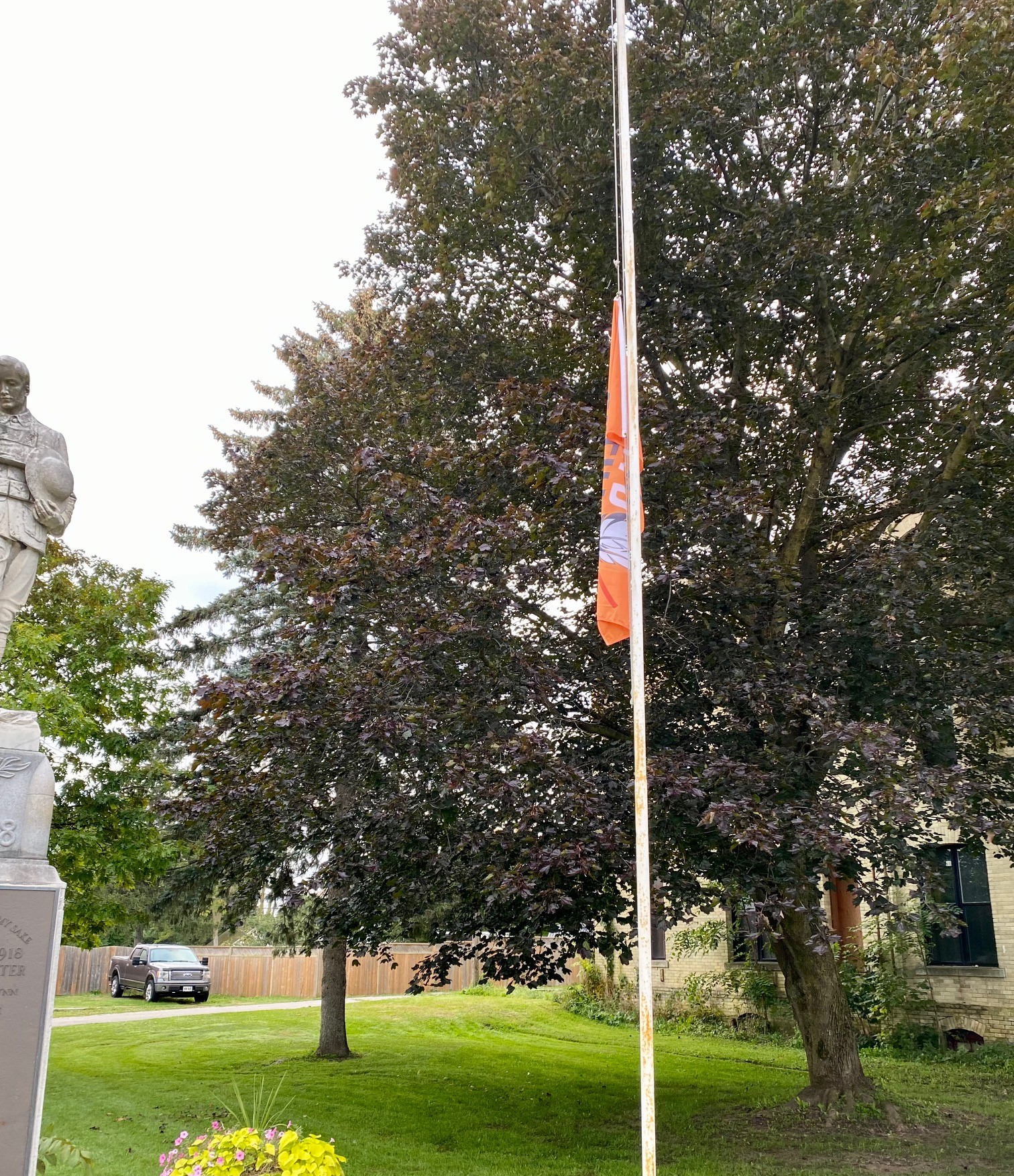 The orange Every Child Matters flag is flown at half mast in the foreground, with a cenotaph statue to the left and a large red maple in the background